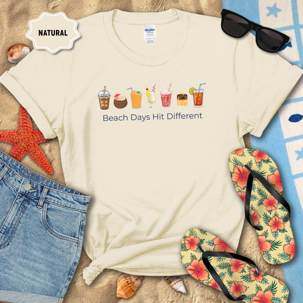 Beach-themed t-shirt with graphics and text, surrounded by flip-flops, sunglasses, and beach accessories on sand.