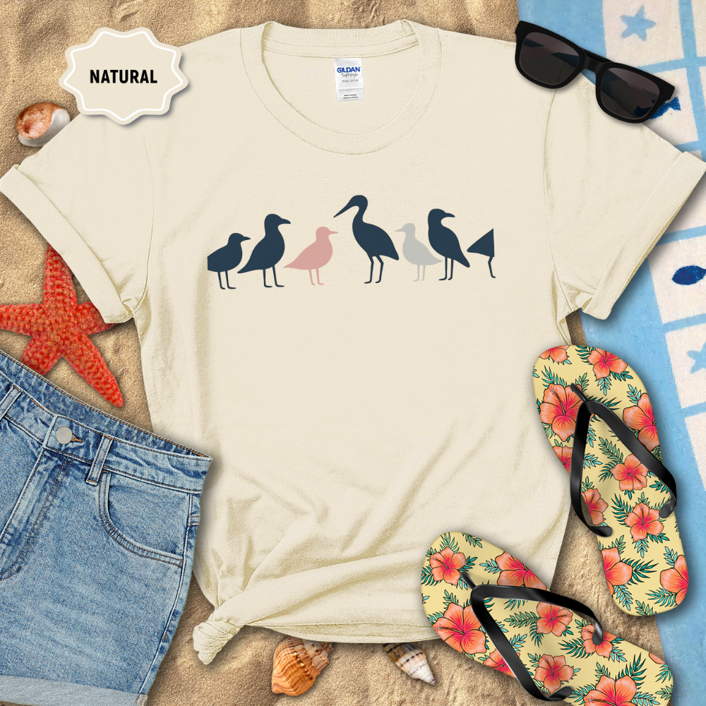Beige t-shirt with bird design on a sandy surface with beach accessories.