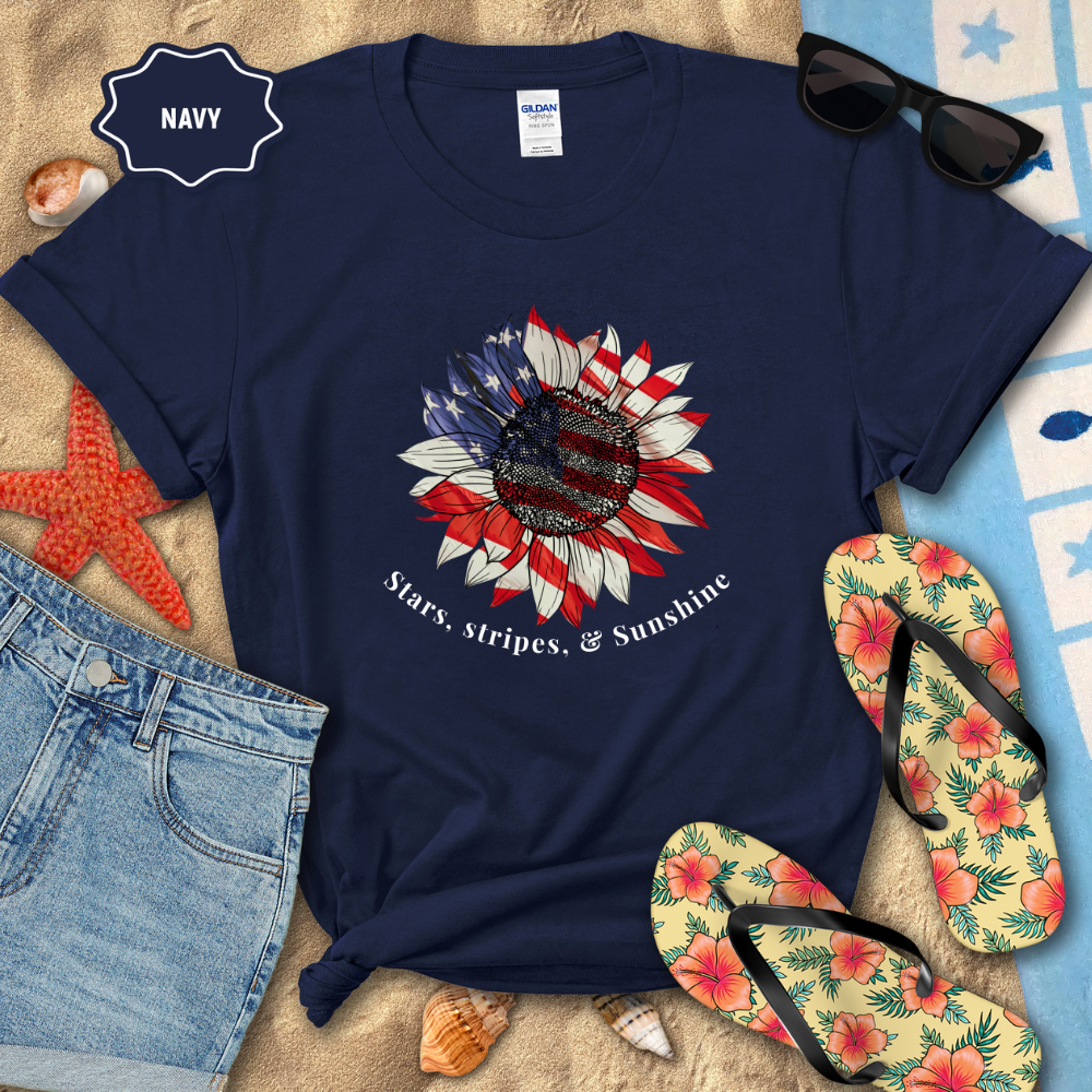 Navy t-shirt with American flag sunflower design, sunglasses, denim shorts, and flip-flops on a beach setting.