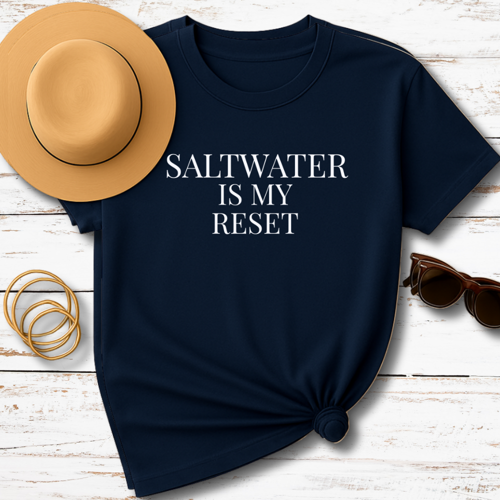 Navy blue t-shirt with 'SALTWATER IS MY RESET' text, tan hat, gold bracelets, and sunglasses on a wooden surface.