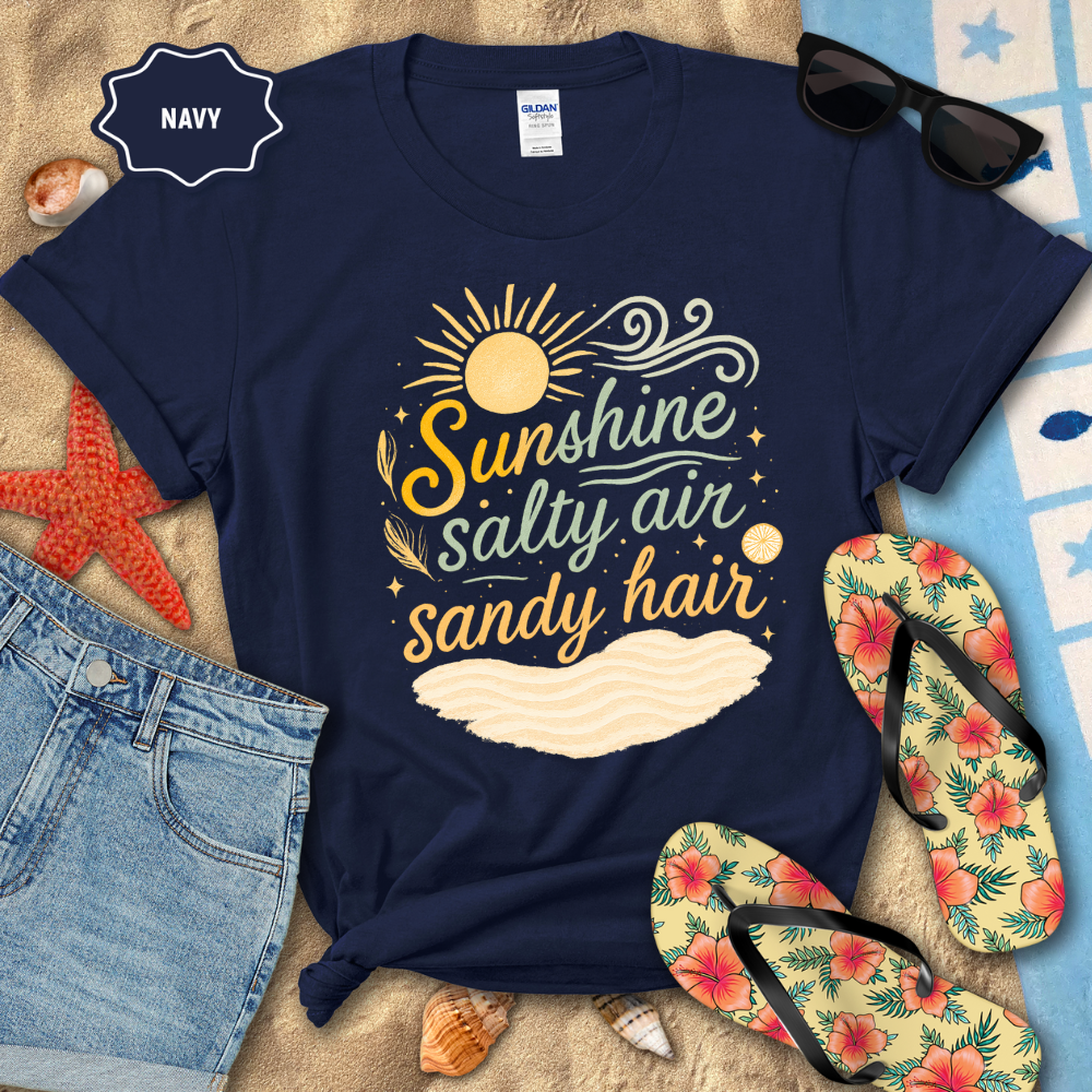 Navy t-shirt with 'Sunshine, Salty Air, Sandy Hair' design on a beach-themed background with sunglasses, flip-flops, and denim shorts.