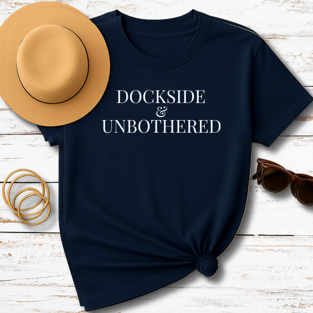 Navy blue t-shirt with 'Dockside & Unbothered' text, tan hat, sunglasses, and gold bracelets on a wooden surface.