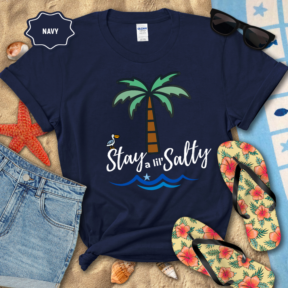 Navy t-shirt with palm tree and 'Stay a l'il' Salty' text, surrounded by beach accessories on sand.
