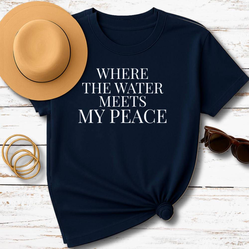 Navy blue t-shirt with 'Where the water meets my peace' text, accompanied by a tan hat, sunglasses, and gold bracelets on a wooden surface.