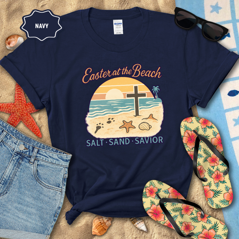 Navy t-shirt with beach-themed design on a sandy surface with flip-flops and sunglasses.