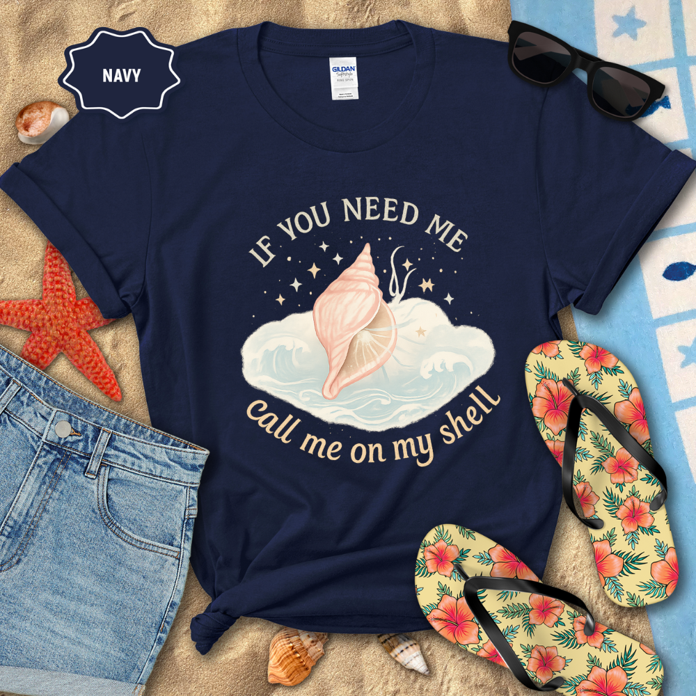 Navy t-shirt with shell graphic and text, surrounded by beach accessories on a sandy surface.