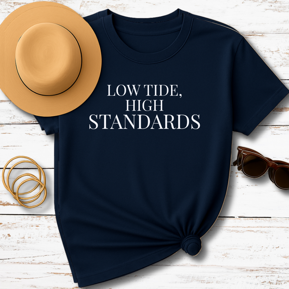 Navy blue t-shirt with 'LOW TIDE, HIGH STANDARDS' text, tan hat, gold bracelets, and sunglasses on a wooden surface.