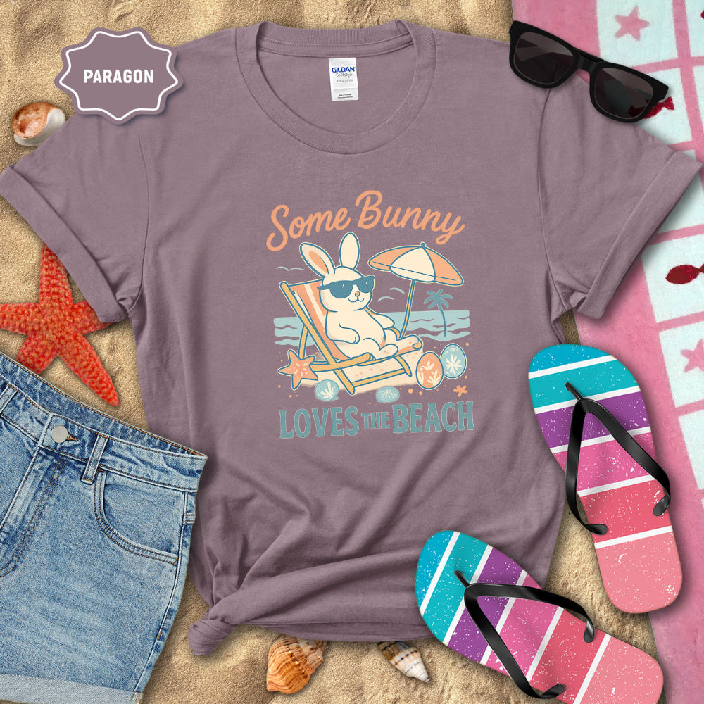 Purple t-shirt with beach-themed design on sand with sunglasses, flip-flops, and denim shorts.