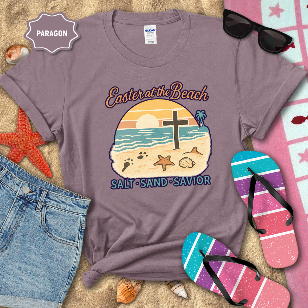 Purple t-shirt with beach-themed design on sand with sunglasses, flip-flops, and denim shorts.