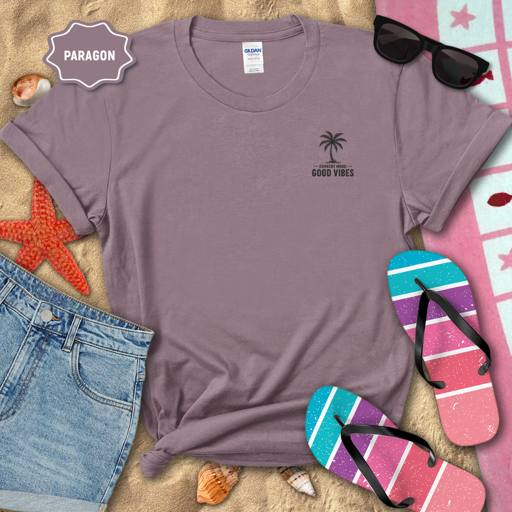 Purple t-shirt with palm tree design and 'Good Vibes' text on a sandy background with beach accessories.