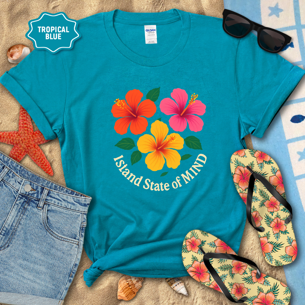 Turquoise t-shirt with tropical flower design and text, surrounded by beach accessories on sand.