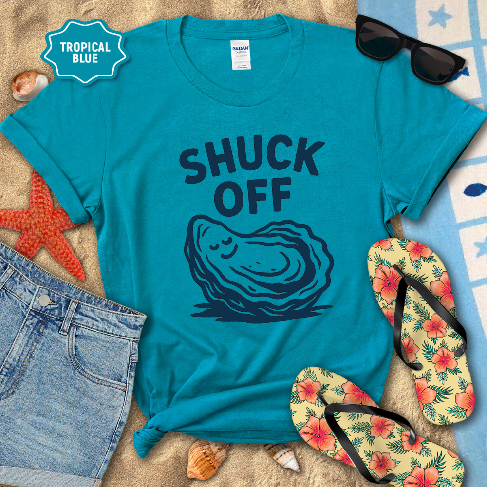 Blue t-shirt with 'SHUCK OFF' graphic and oyster illustration, surrounded by beach accessories on sand.