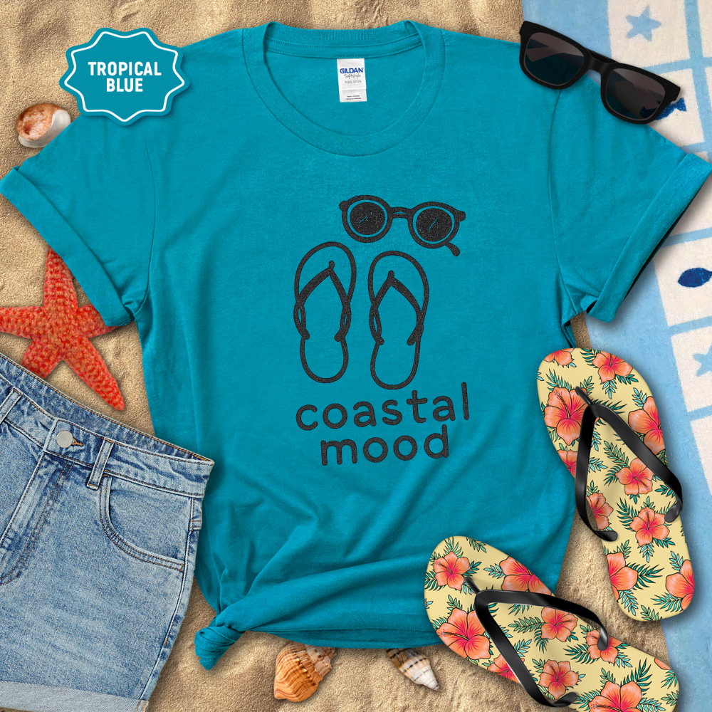 Blue t-shirt with 'coastal mood' design, sunglasses, flip-flops, and beach items on a sandy background.