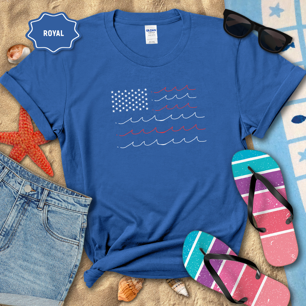 Blue t-shirt with American flag design on a sandy surface with beach accessories.