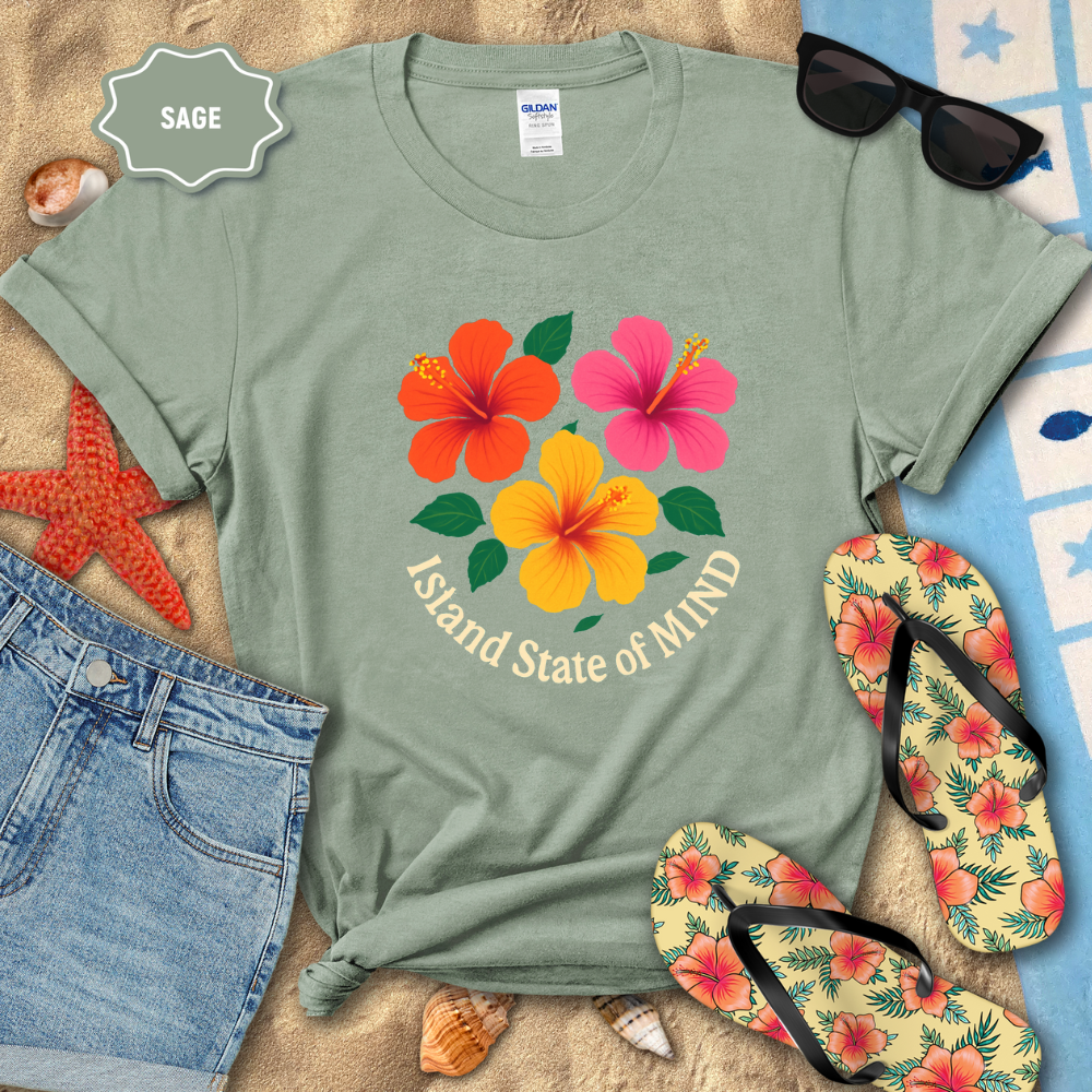 Sage green t-shirt with floral design and text, surrounded by beach accessories on sand.