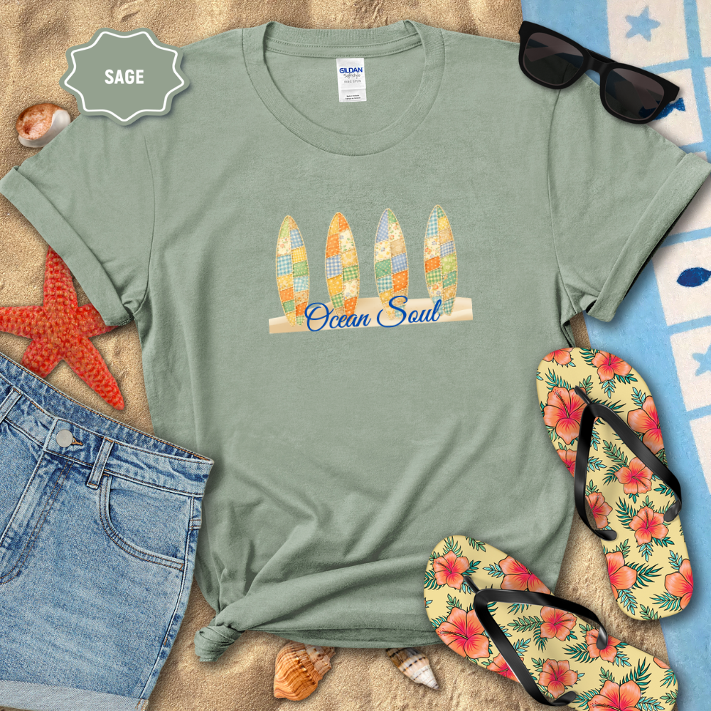 Sage green t-shirt with 'Ocean Soul' design on a sandy surface with beach accessories.