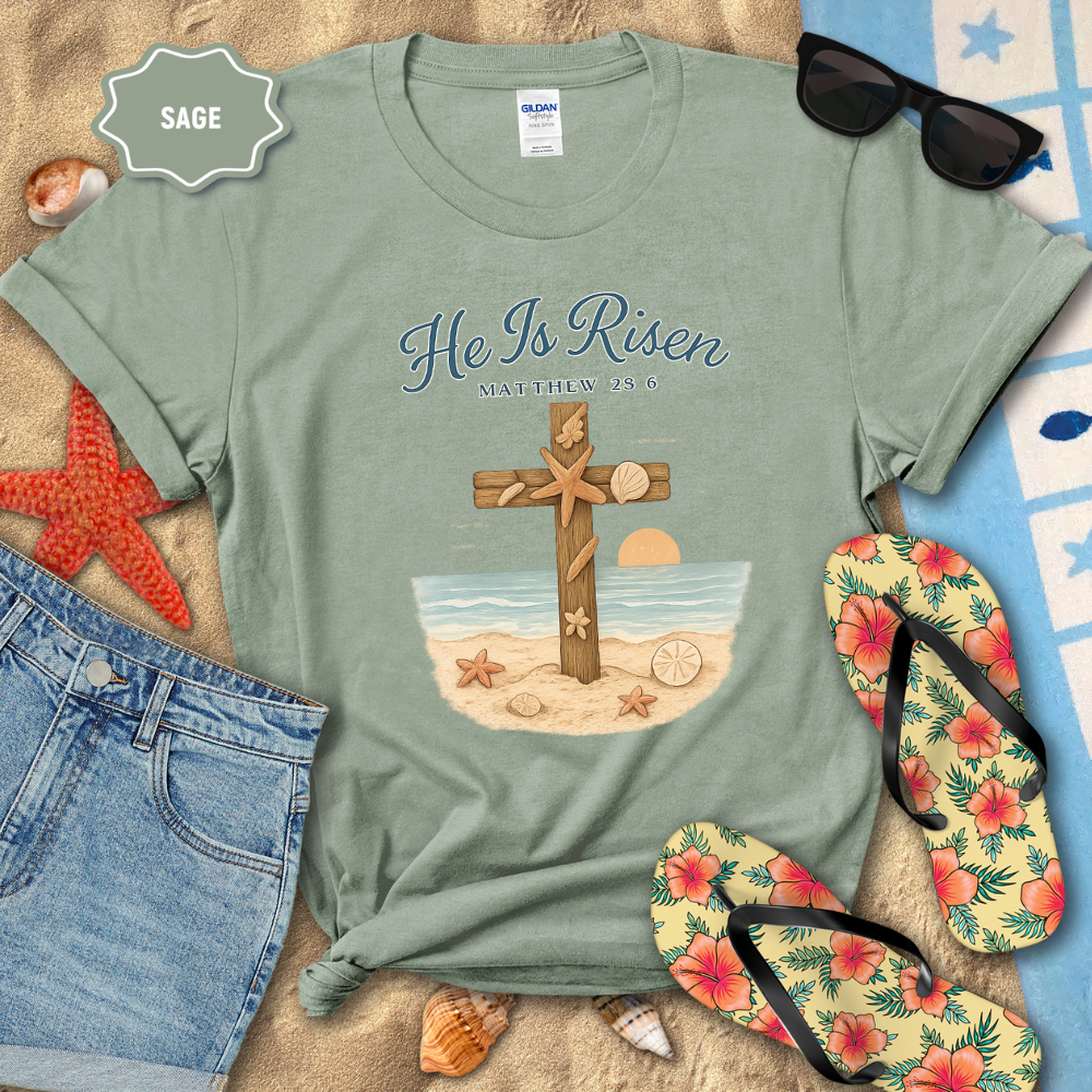 Sage green t-shirt with 'He Is Risen' design on a sandy surface with beach accessories.