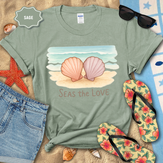 Sage green t-shirt with beach-themed design featuring two shells and 'Seas the Love' text, surrounded by beach accessories.