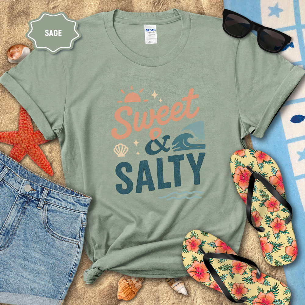 Sage green t-shirt with 'Sweet & Salty' text, sunglasses, flip-flops, and beach items on sand.