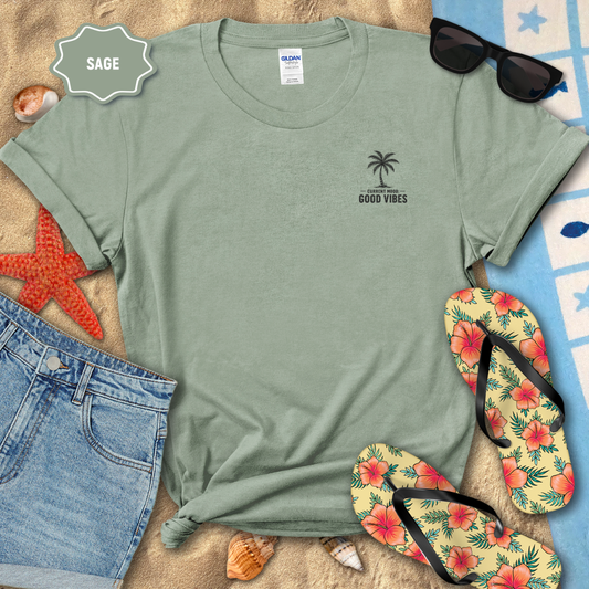 Sage green t-shirt with palm tree design and 'Good Vibes' text, surrounded by beach accessories on sand.