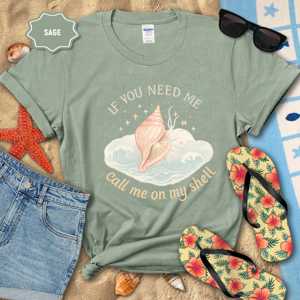 Sage green t-shirt with beach-themed design on a sandy surface with flip-flops and sunglasses.