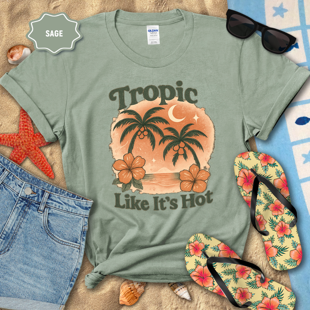 Sage green t-shirt with tropical design and text 'Tropic Like It's Hot' on a beach-themed background.