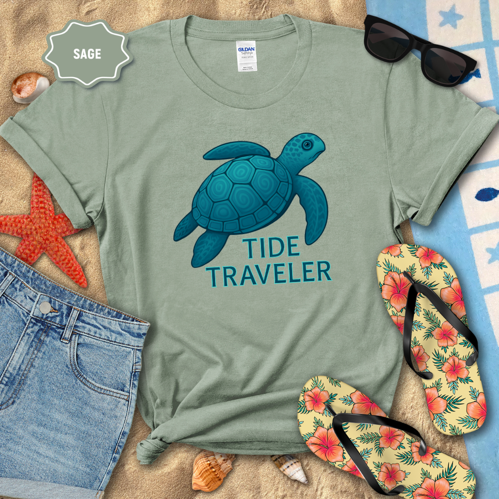 Sage green t-shirt with blue turtle graphic and 'Tide Traveler' text, surrounded by beach accessories.