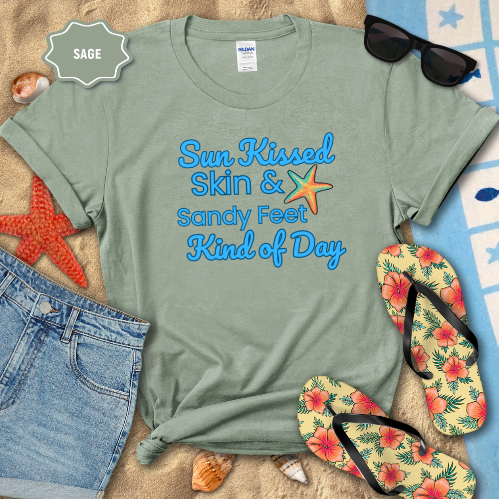 Sage green t-shirt with 'Sun Kissed Skin & Sandy Feet Kind of Day' text, sunglasses, flip-flops, and beach accessories on sand.