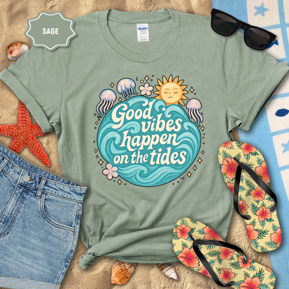 Sage green t-shirt with 'Good vibes happen on the tides' design, sunglasses, flip-flops, and beach items on sand.