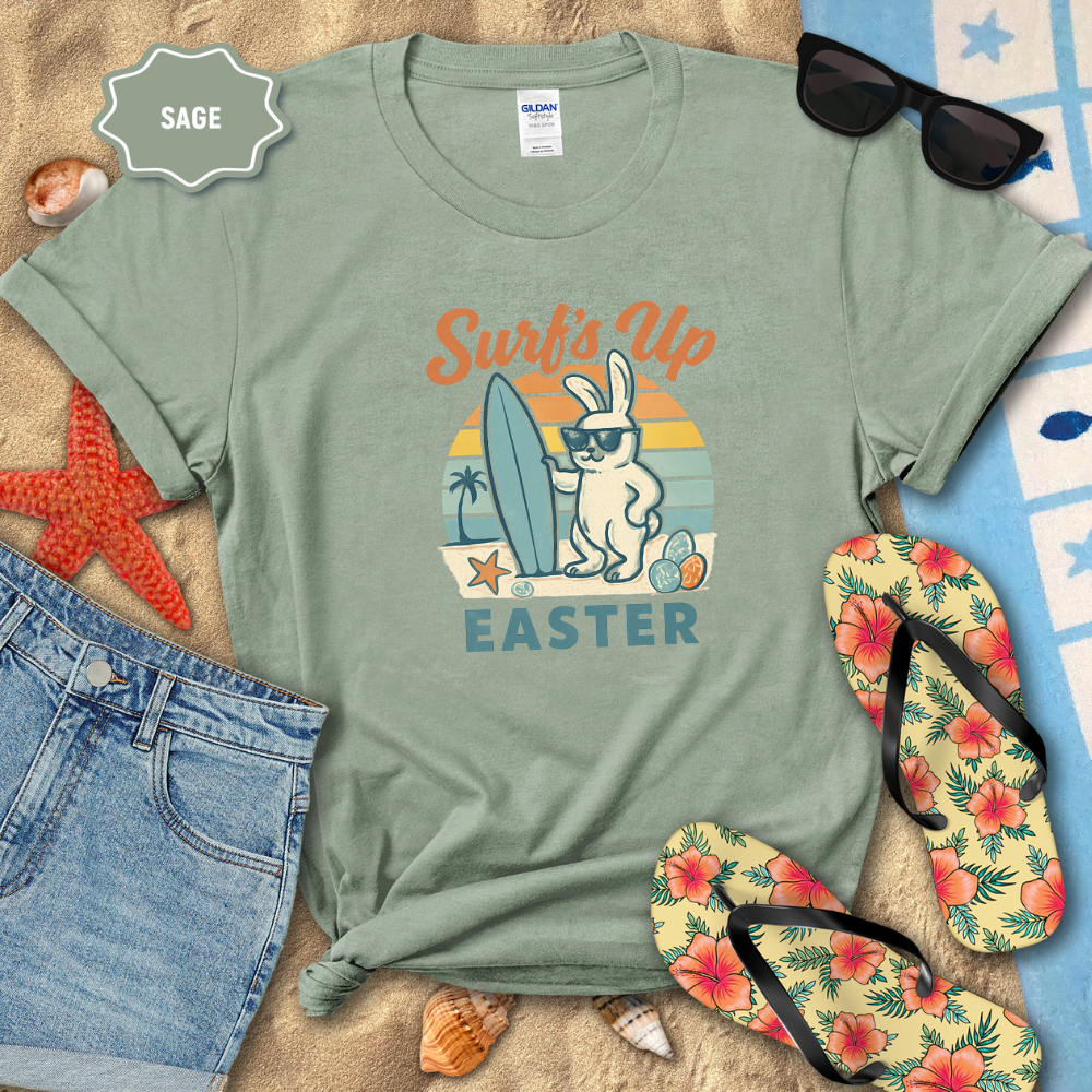 Sage green t-shirt with Easter bunny design on a sandy surface with flip-flops and sunglasses.