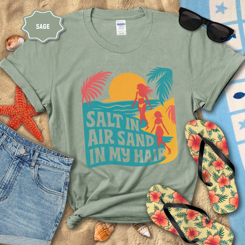 Sage green t-shirt with beach graphic and text 'Salt in air, sand in my hair' on a sandy background with flip-flops and sunglasses.