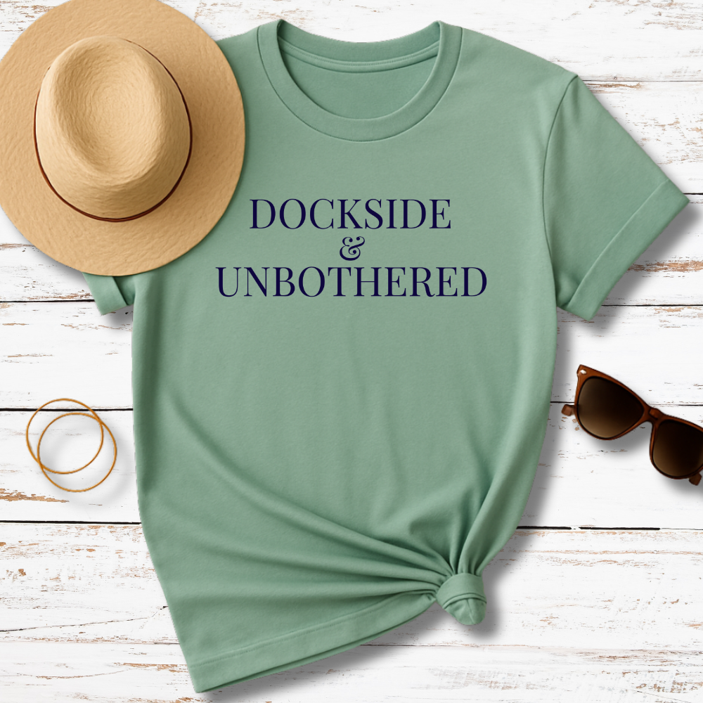 Green t-shirt with 'Dockside & Unbothered' text, accompanied by a hat, sunglasses, and bracelet on a wooden surface.