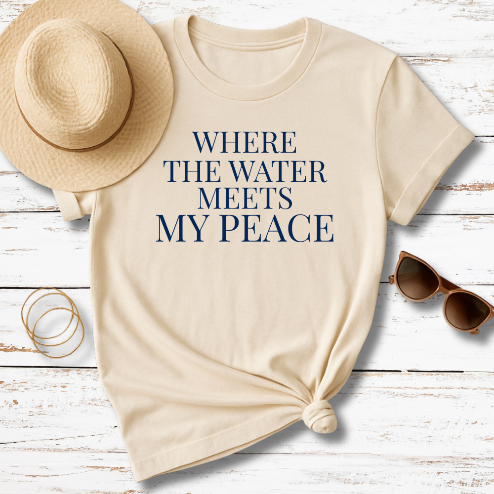 Beige t-shirt with 'WHERE THE WATER MEETS MY PEACE' text, sun hat, sunglasses, and bracelet on a wooden surface