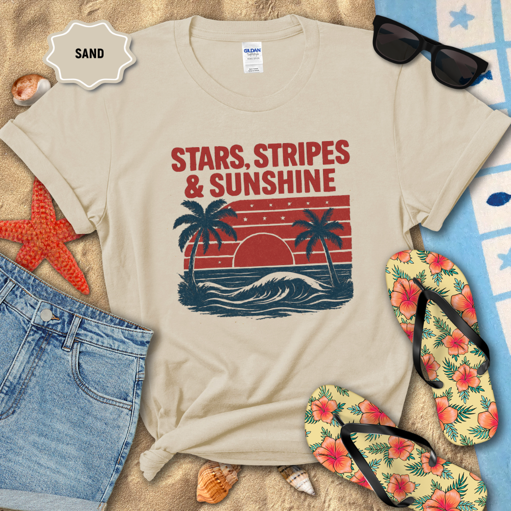 Beige t-shirt with 'Stars, Stripes & Sunshine' design on sand with beach accessories.