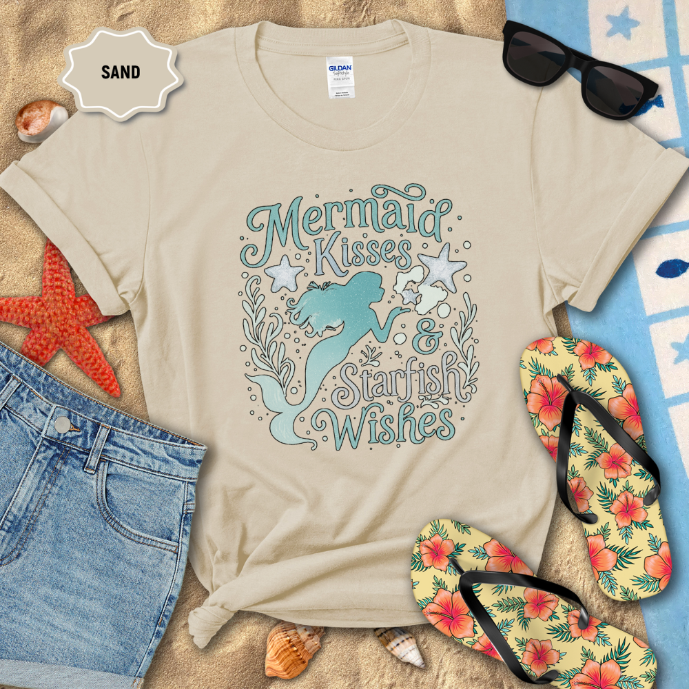 Beige t-shirt with mermaid design on sand with sunglasses, flip-flops, and beach accessories.