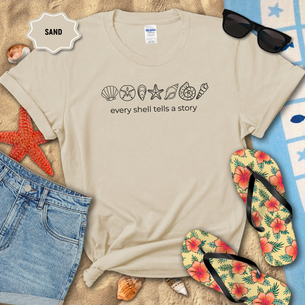 Beige t-shirt with beach-themed design and text, surrounded by sand, flip-flops, and shells.
