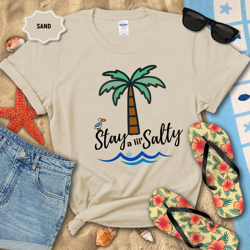 Beige t-shirt with palm tree and 'Stay a lil' Salty' text on sand with beach accessories.