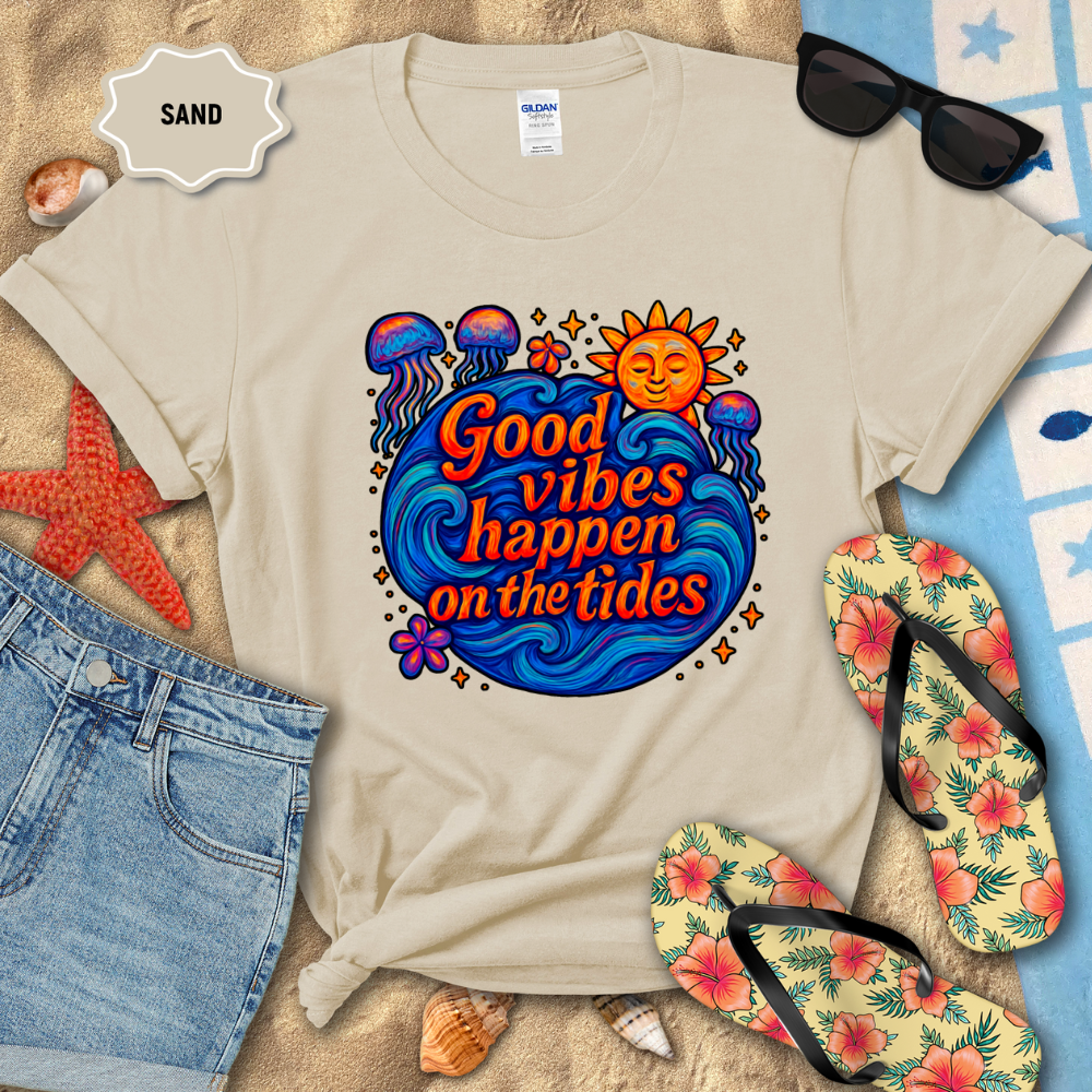 Beige t-shirt with 'Good vibes happen on the tides' design on a sandy background with beach accessories.