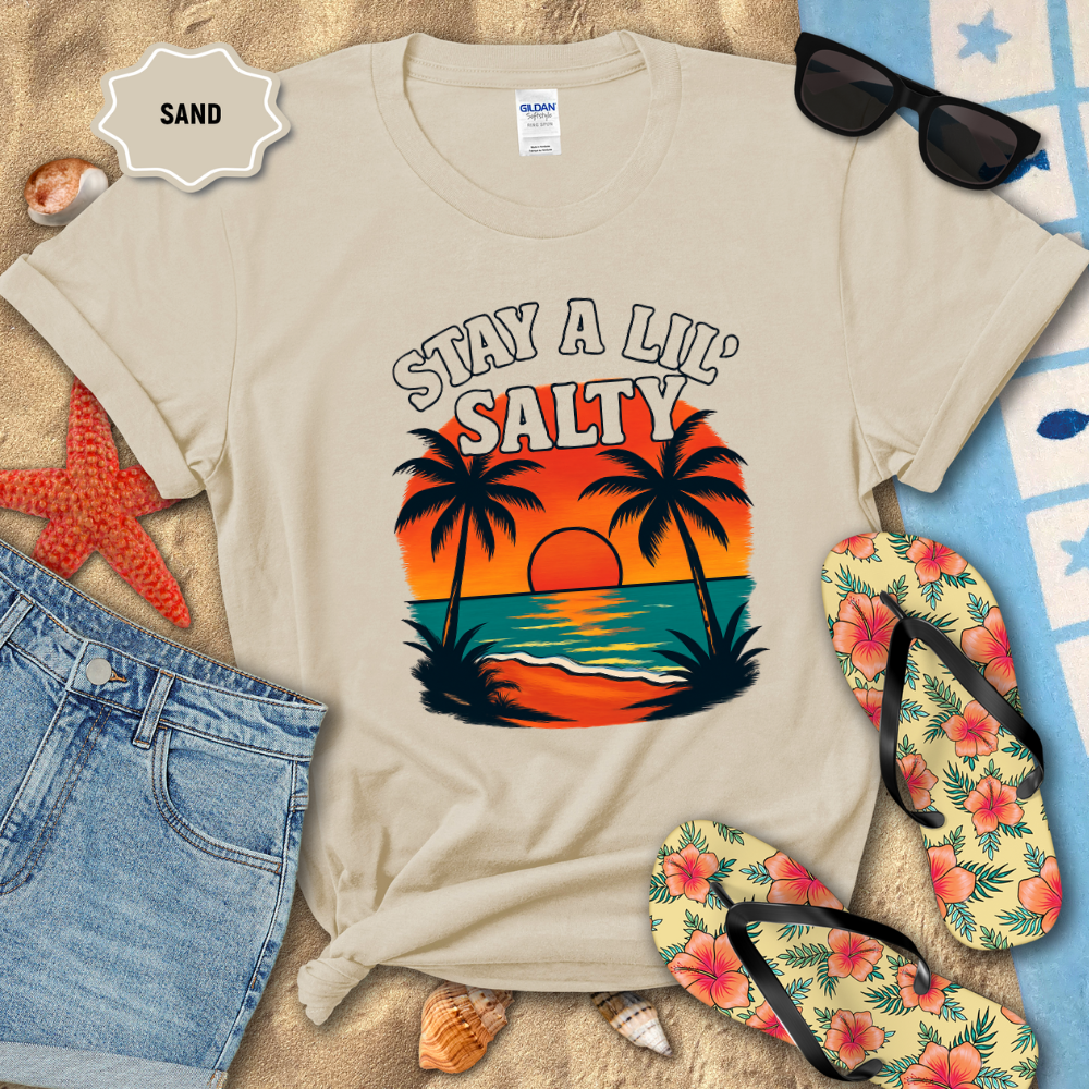 Beige t-shirt with 'Stay a Lil' Salty' design on sand with beach accessories.
