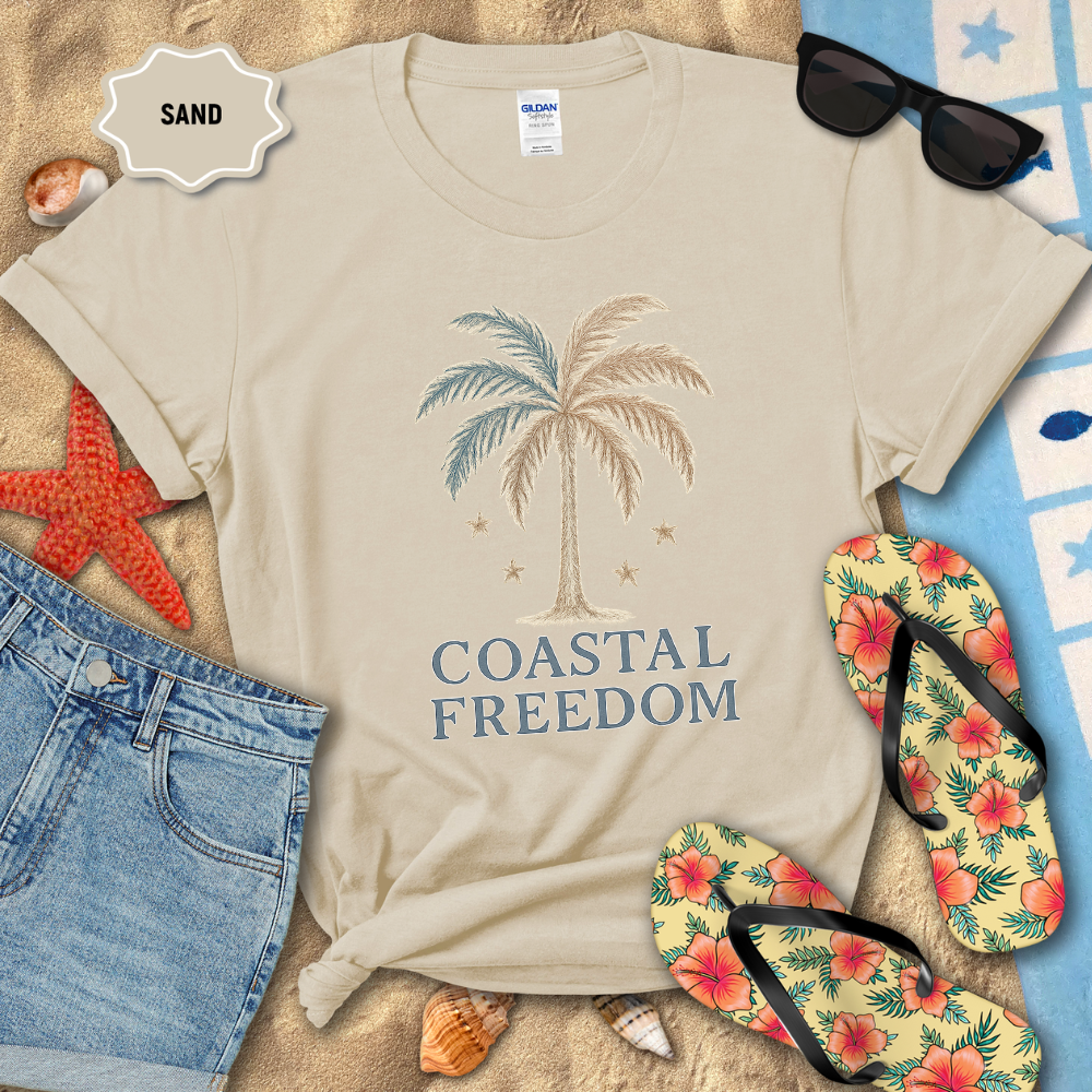 Beige t-shirt with 'Coastal Freedom' design on a sandy background with beach accessories.