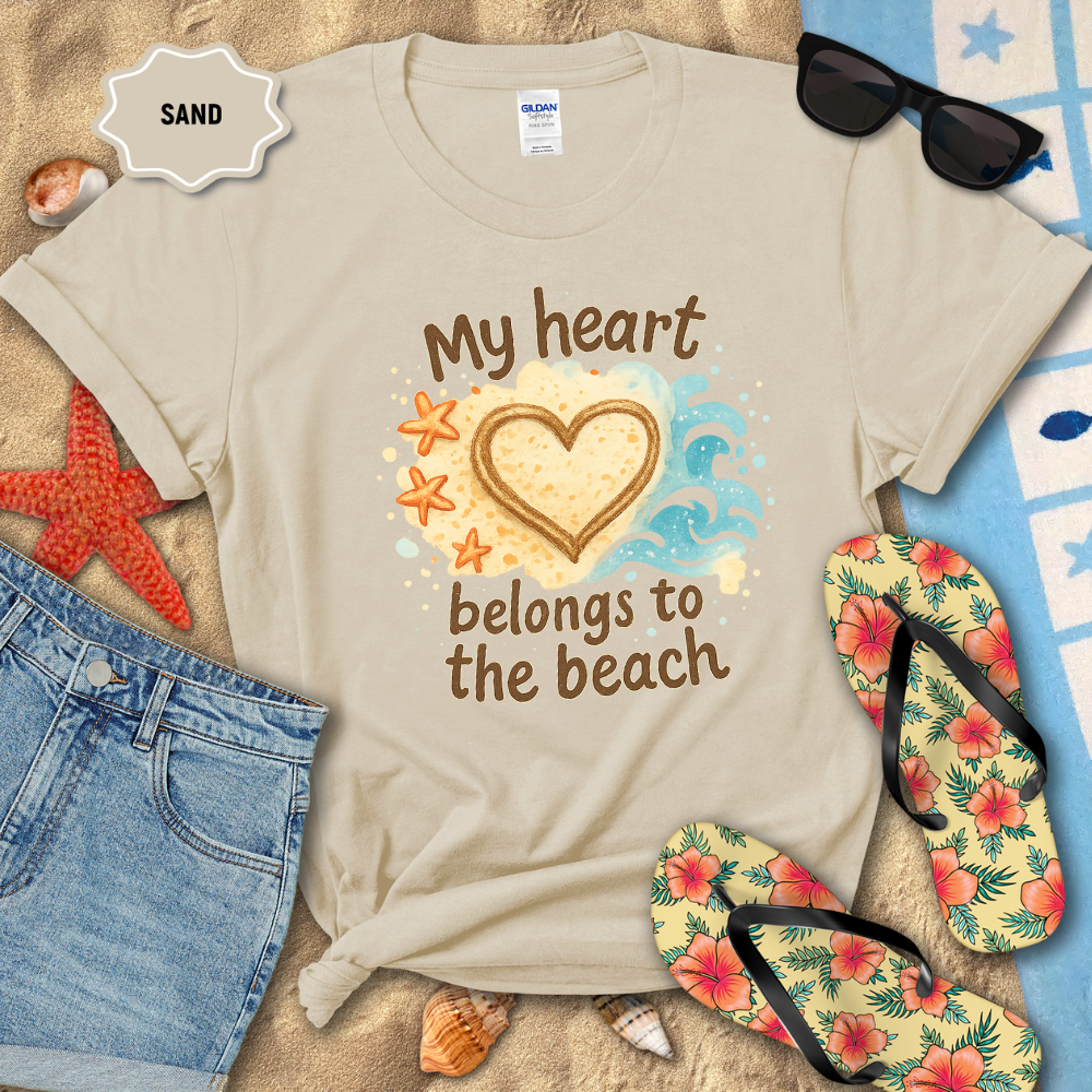 Beige t-shirt with 'My heart belongs to the beach' design on sand with sunglasses, flip-flops, and beach accessories.
