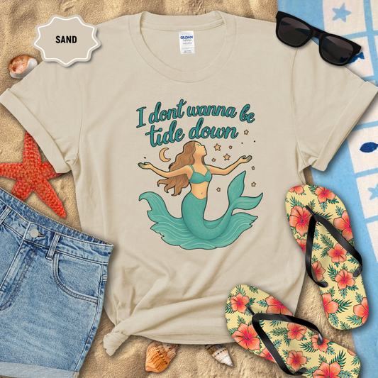 Beige t-shirt with mermaid graphic and text on sand with beach accessories.