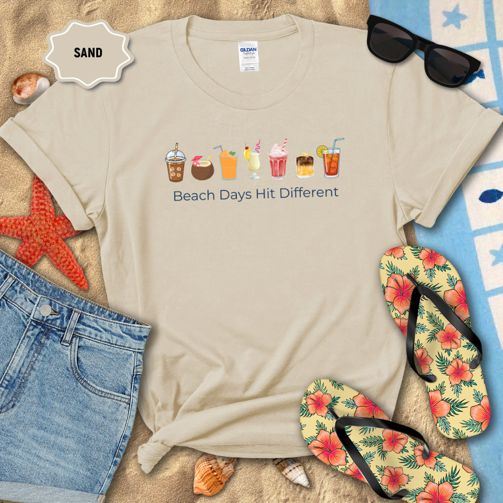 Beige t-shirt with beach-themed graphics and text, surrounded by beach accessories on sand.