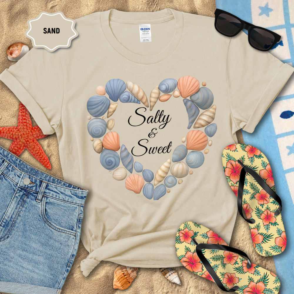 Beige t-shirt with shell design and 'Salty & Sweet' text, surrounded by beach accessories on sand.