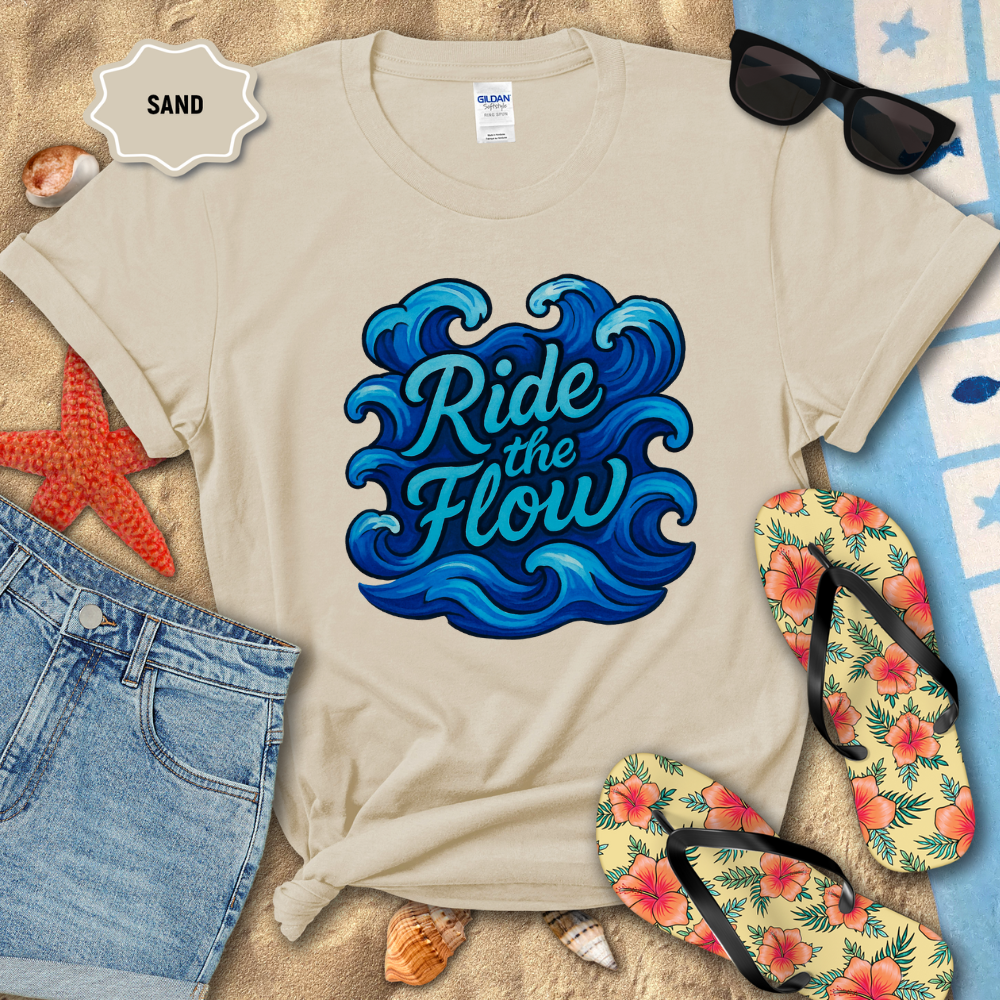 Beige t-shirt with 'Ride the Flow' design on sand, surrounded by beach accessories.