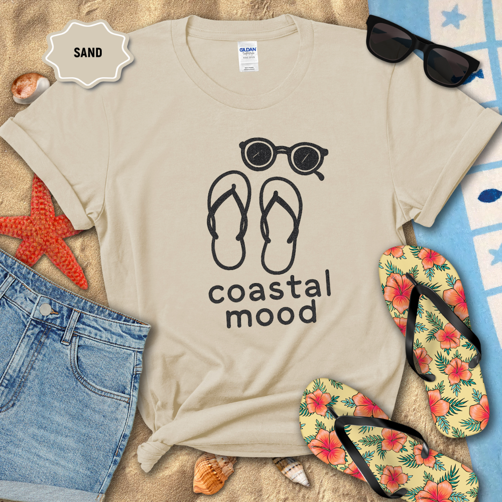 Beige t-shirt with 'coastal mood' design on sand with beach accessories.