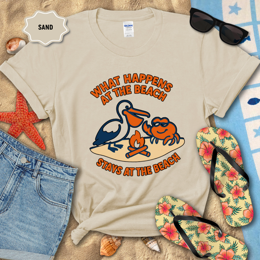 Beige t-shirt with beach-themed design featuring a pelican and crab, surrounded by beach accessories on sand.