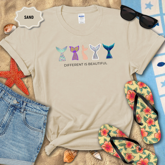 Beige t-shirt with mermaid tail designs and 'Different is Beautiful' text on a sandy background with beach accessories.