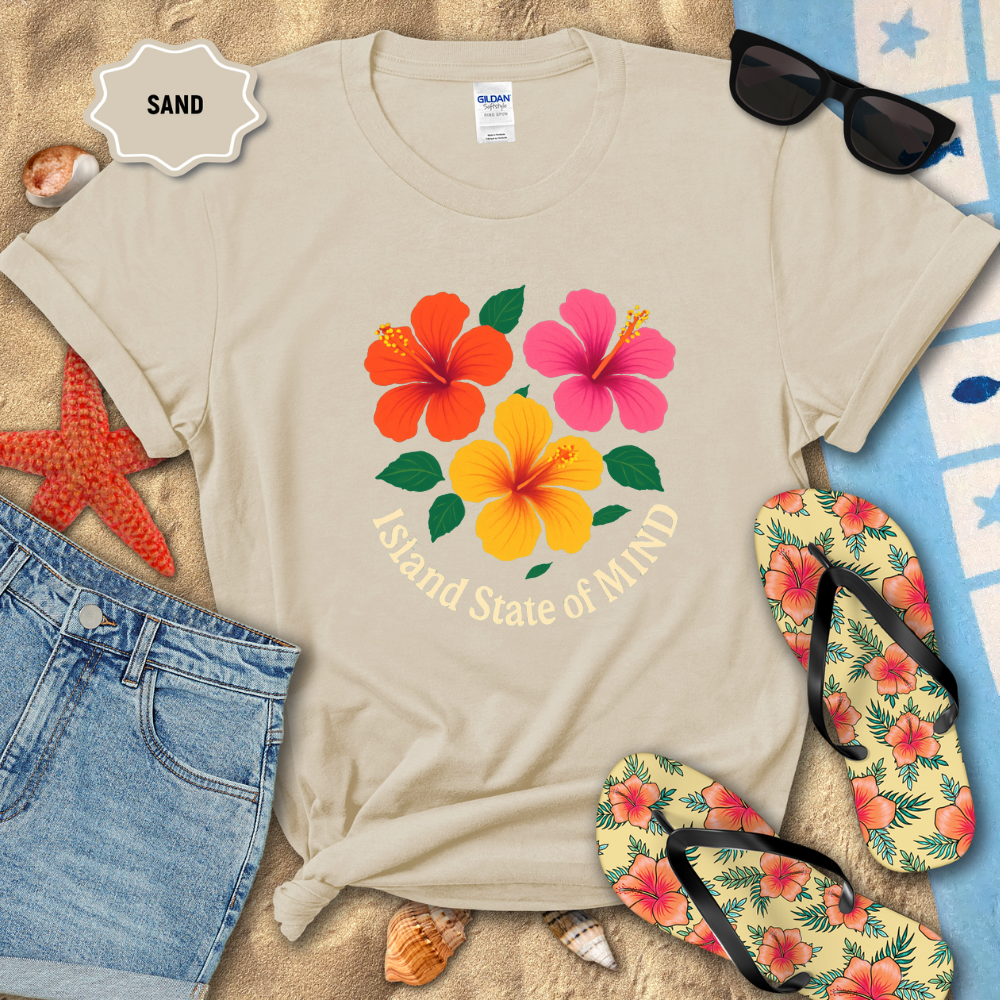 Beige t-shirt with floral design and text on a sandy background with beach accessories.