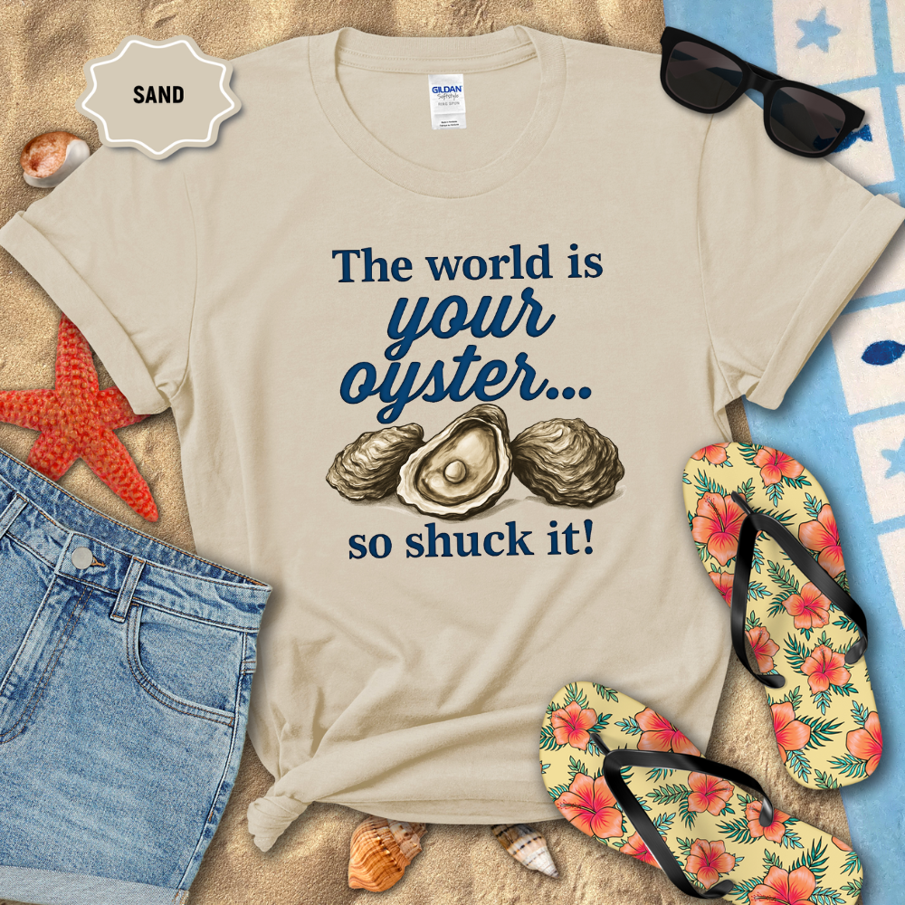 Beige t-shirt with oyster graphic and text on a sandy background with beach accessories.
